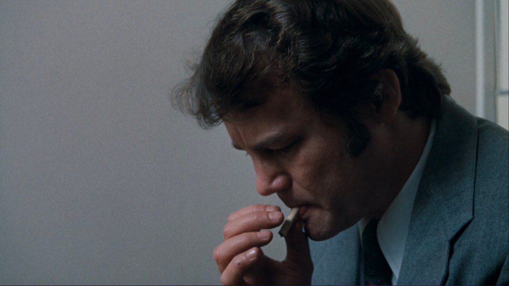 Close-up of a man in a suit, looking down thoughtfully while holding a cigarette near his mouth.