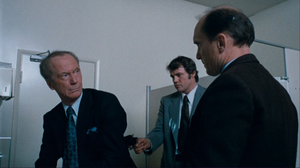 Three men in a bathroom setting, with one man in a suit appearing to react to another man who is holding a gun.