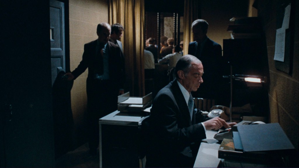 A man in a suit sits at a desk, using a typewriter, while several people are gathered in the background. The scene is dimly lit, with a warm glow from a desk lamp.