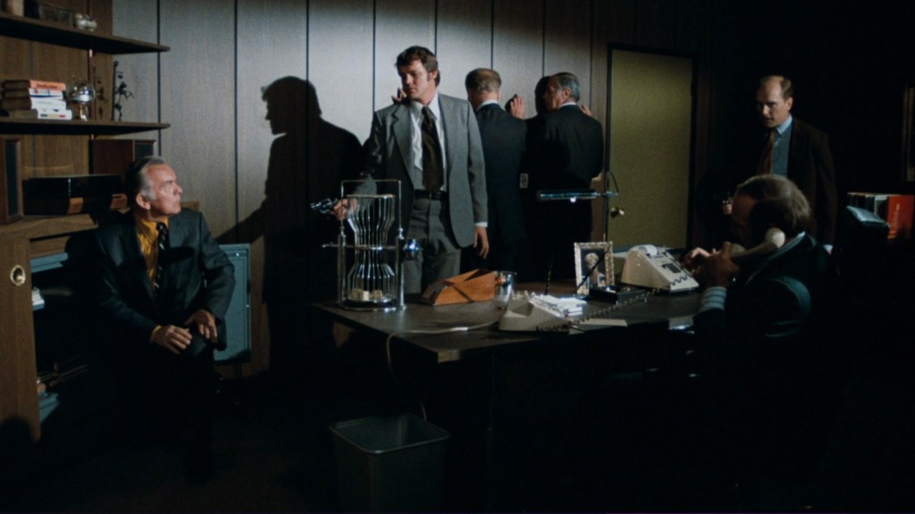 A tense office scene with six men in suits, one seated and the others standing or leaning against the wall, engaged in a serious conversation. A cluttered desk with a telephone, a framed photo, and a birdcage is visible.