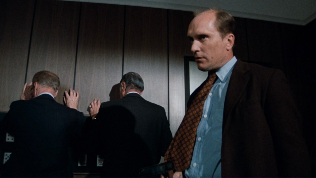A tense scene featuring a man holding a gun, standing close to two other men who have their backs turned against a wall.