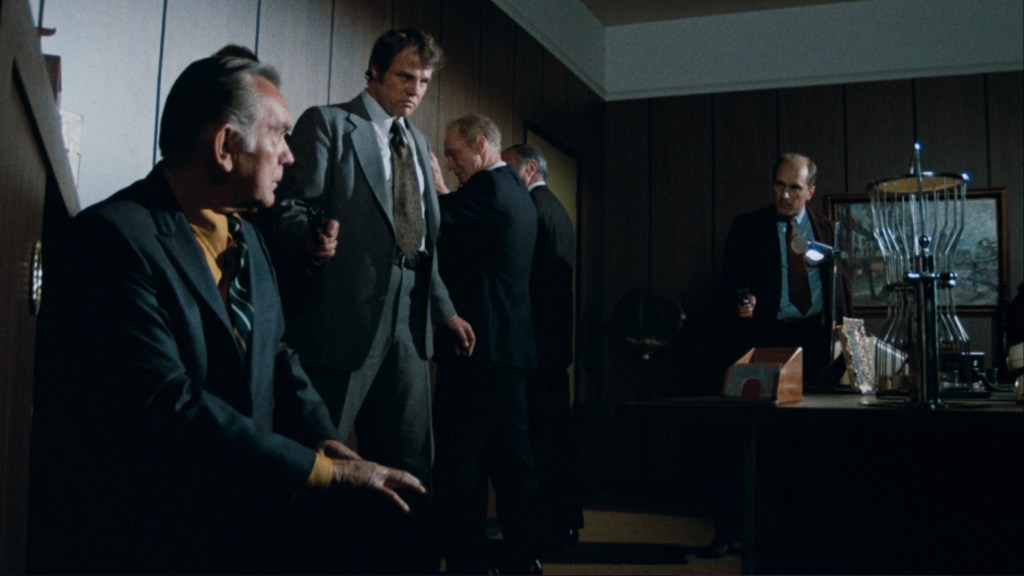 A tense scene in an office with four men, two standing and two sitting, as one holds a gun and another appears to be searching for something.
