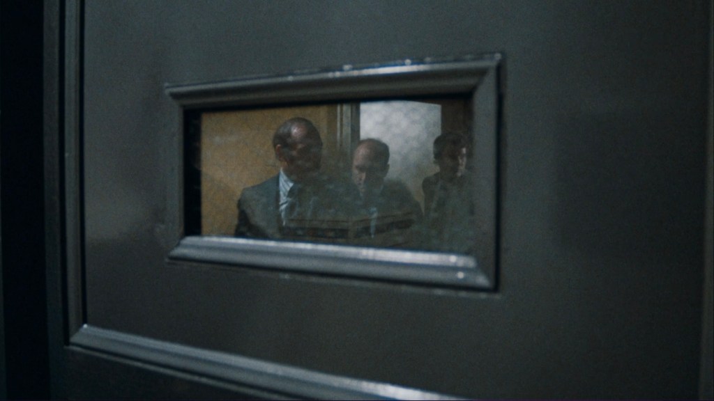 Three men are seen through a small window in a door, appearing to be in a serious discussion.
