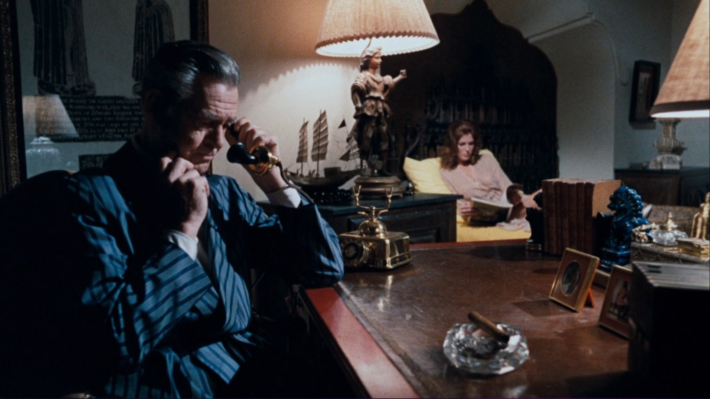 A man in a striped suit sits at a desk with a worried expression, holding a telephone to his ear. In the background, a woman in a long dress reads a book while lounging on a yellow chair, surrounded by decorative items and lamps.