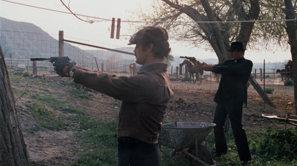 Two men in a rural setting are aiming handguns at each other, with a scenic background featuring trees and mountains.