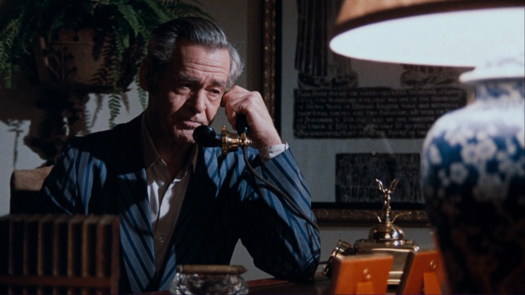 A man in a striped suit sitting at a desk, holding a telephone to his ear with a thoughtful expression. A lamp and decorative items are visible on the desk, along with a patterned vase and a wall with framed art.