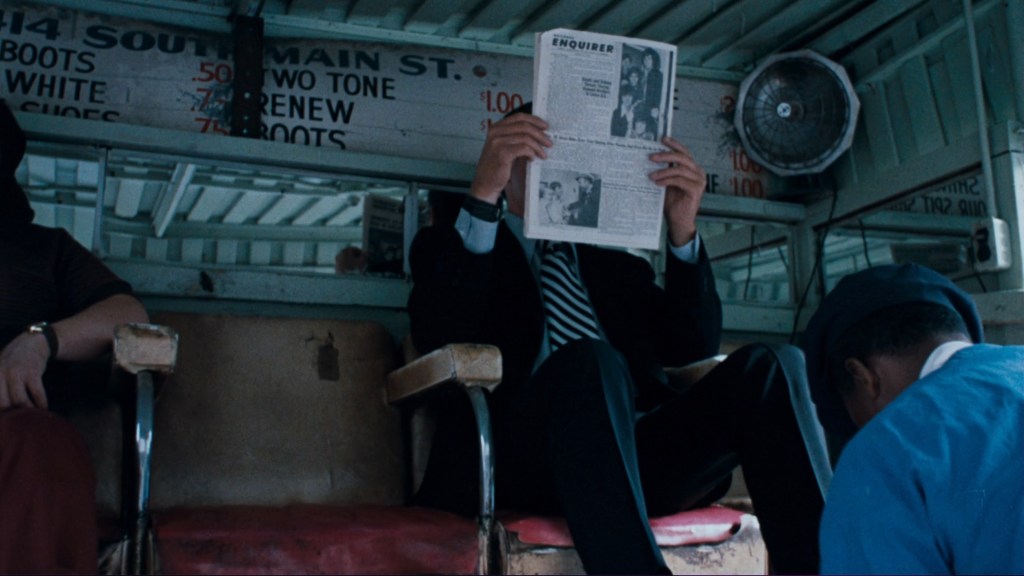 A man in a suit reading a newspaper while sitting on a bus, with another man seated nearby and a worker attending to the bus interior.
