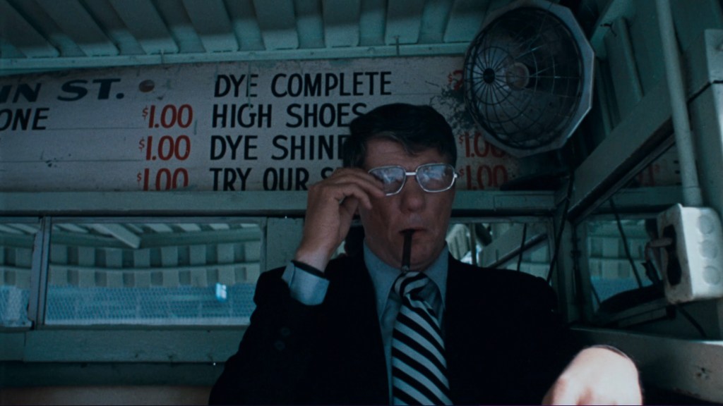 A man in a suit and glasses sitting in a booth, holding a pen to his mouth, with a sign displaying prices for shoe dyeing services in the background.
