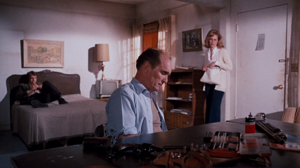 A tense scene in a dimly lit room featuring a man sitting at a table with various items, a woman standing nearby, and another man lounging on a bed in the background.