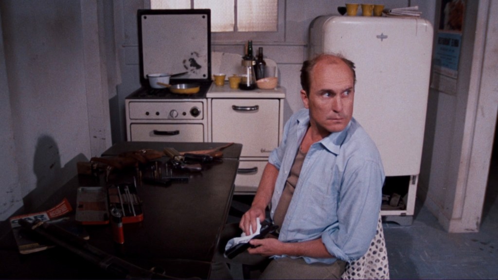 A man seated at a table in a dimly lit kitchen, holding a firearm and looking over his shoulder. The kitchen features vintage appliances and scattered tools on the table.