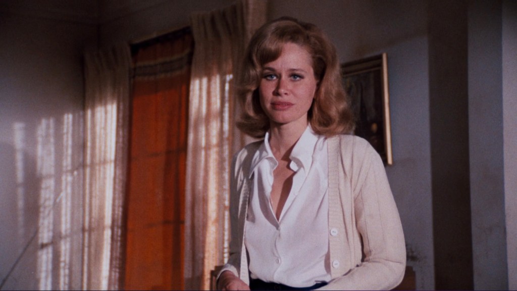 A woman with blonde wavy hair is standing in a room with soft lighting. She is wearing a white blouse and a light cardigan, presenting a serious expression.