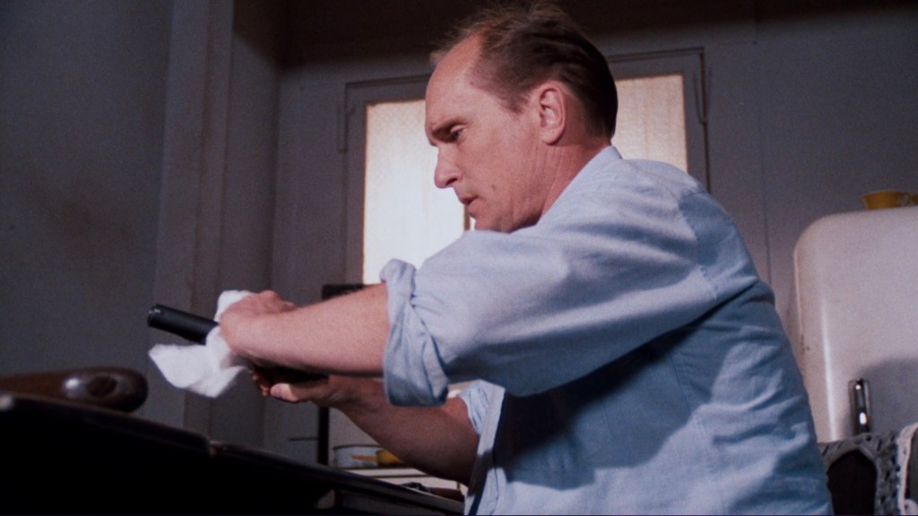 A man in a light blue shirt cleans a handgun with a white cloth while seated at a table in a domestic setting.
