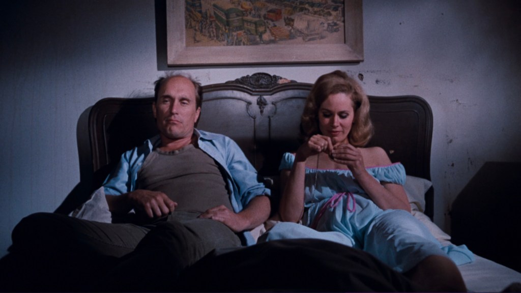 A man and a woman sitting on a bed in a dimly lit room, with the man looking contemplative and the woman engaged in an activity.