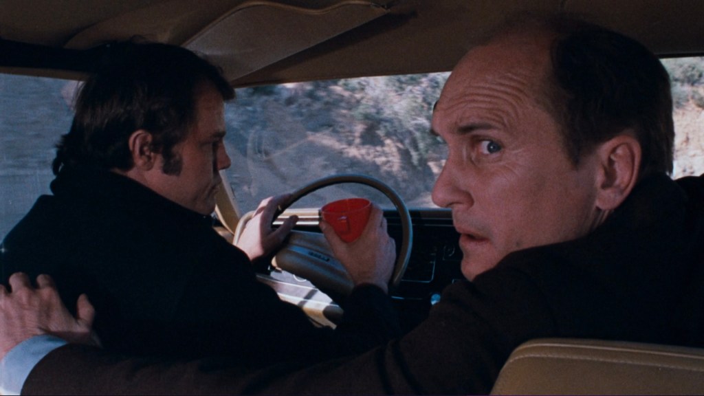 Two men in a car, one driving while the other looks back nervously, holding a red cup.