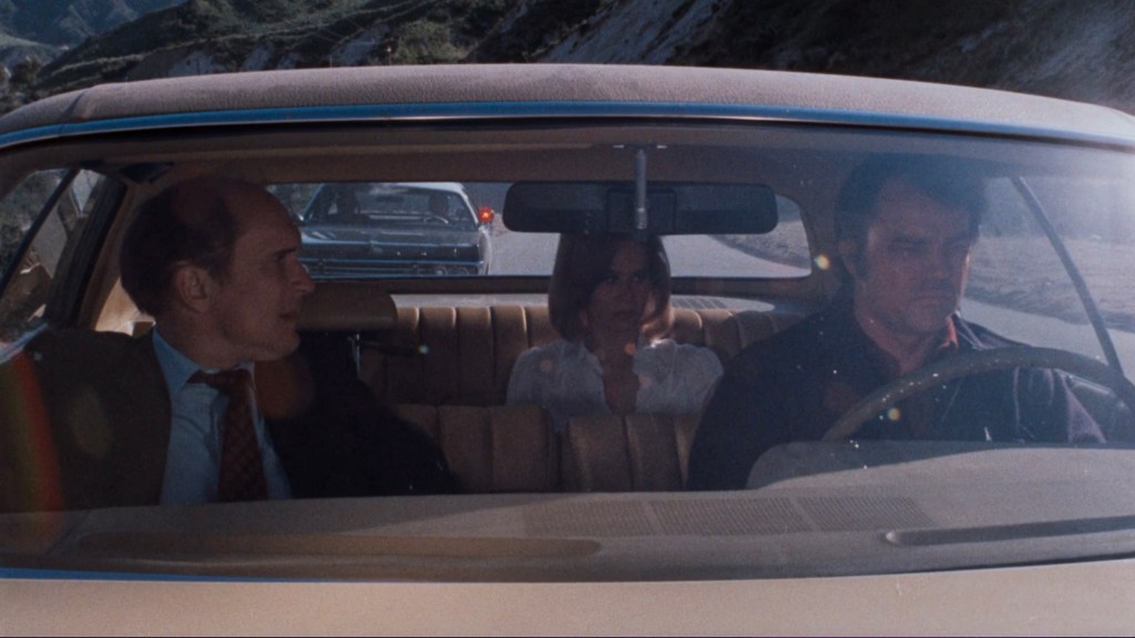 Interior view of a car with three people: a man driving, a woman sitting in the middle, and another man in the passenger seat; scenic mountain road visible outside the windshield.