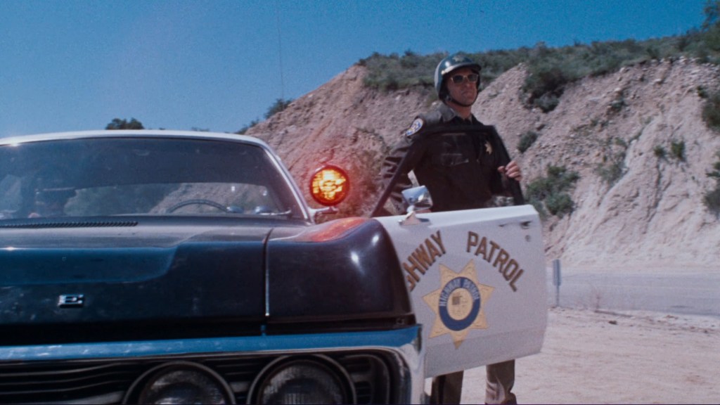 A highway patrol officer stands beside a vintage black and white police car with flashing lights, set against a backdrop of a rugged hillside.
