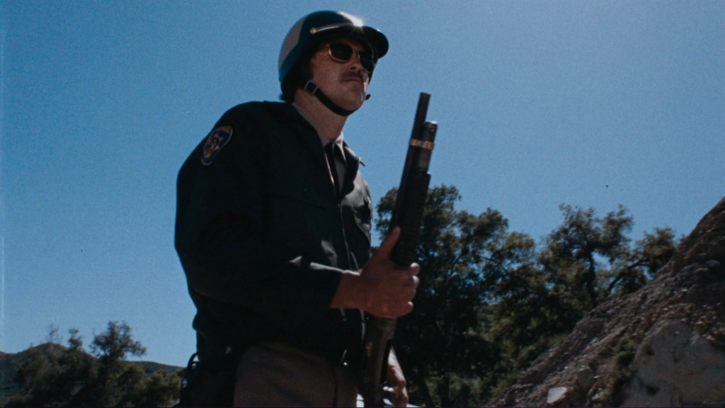 A police officer wearing a helmet and sunglasses holds a firearm while standing outdoors under a clear blue sky.