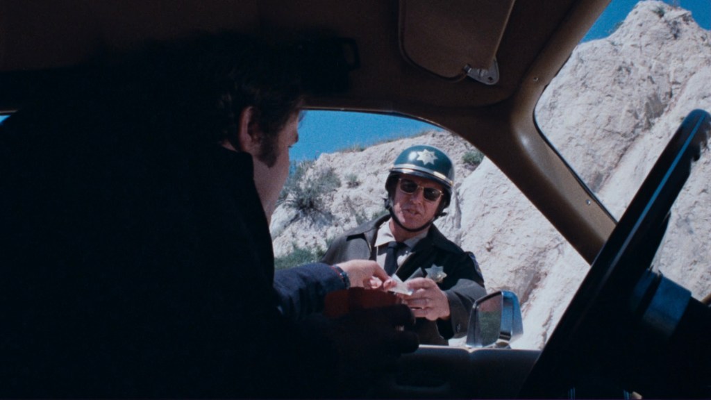 A police officer in a green uniform and sunglasses interacts with a man sitting in a car, with rocky terrain in the background.