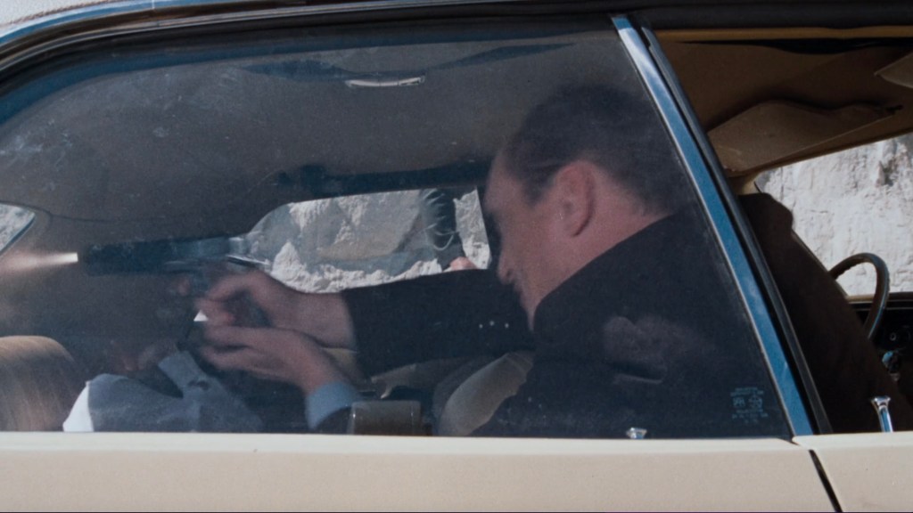 A close-up view of a man inside a car, holding a gun while interacting with another person in the driver's seat, capturing an intense moment.