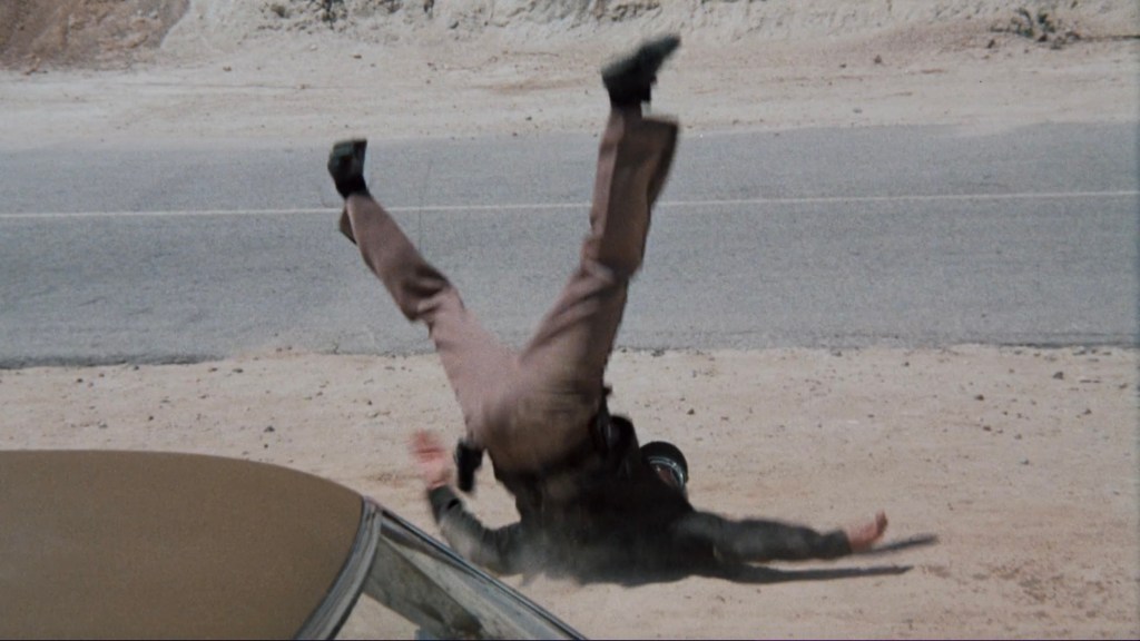 A person falling backwards onto the ground near a roadside, with their legs in the air and a car visible in the foreground.