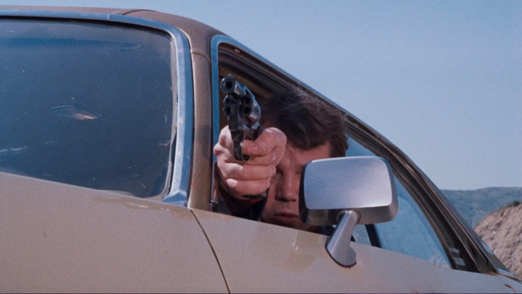A person aiming a handgun from the window of a car with a mountain backdrop.
