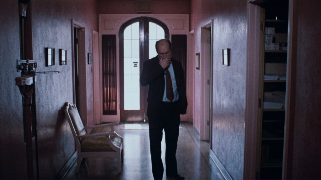A man in a suit stands in a hallway, looking contemplatively at the floor; the walls are painted pink, and there are framed pictures on the walls.