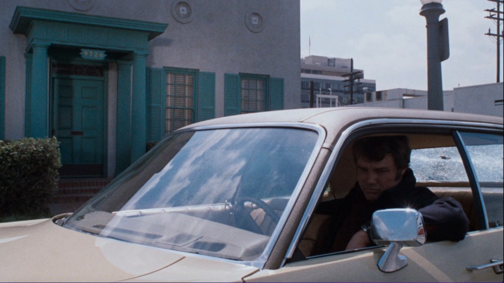 A man sitting in a vintage car parked in front of a teal door with shutters. He appears pensive, looking down, with urban buildings visible in the background.