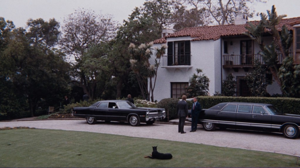Two men in suits standing near black cars in front of a house with palm trees and a dog lying on the lawn.