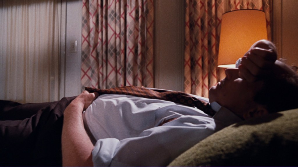 A man lying on a bed, looking distressed, with his hand on his forehead and wearing a dress shirt and tie. Soft lighting from a nearby lamp and patterned curtains in the background.