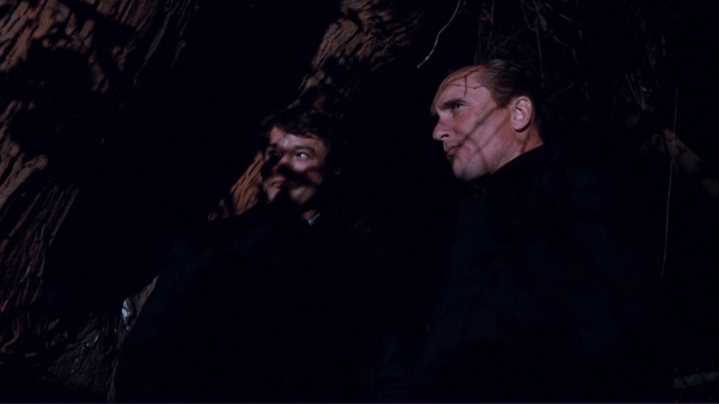 Two men standing close together in a dimly lit forest setting, with shadows of tree branches cast across their faces.