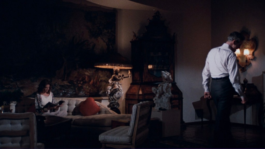 A woman sitting on a couch reading a magazine, with a lamp illuminating the scene, while a man walks away in a dimly lit room filled with decorative furniture.