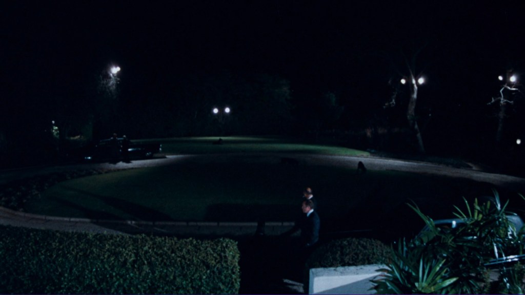 A dimly lit garden at night with two figures walking on a path, surrounded by manicured hedges and faintly illuminated by distant lamps.