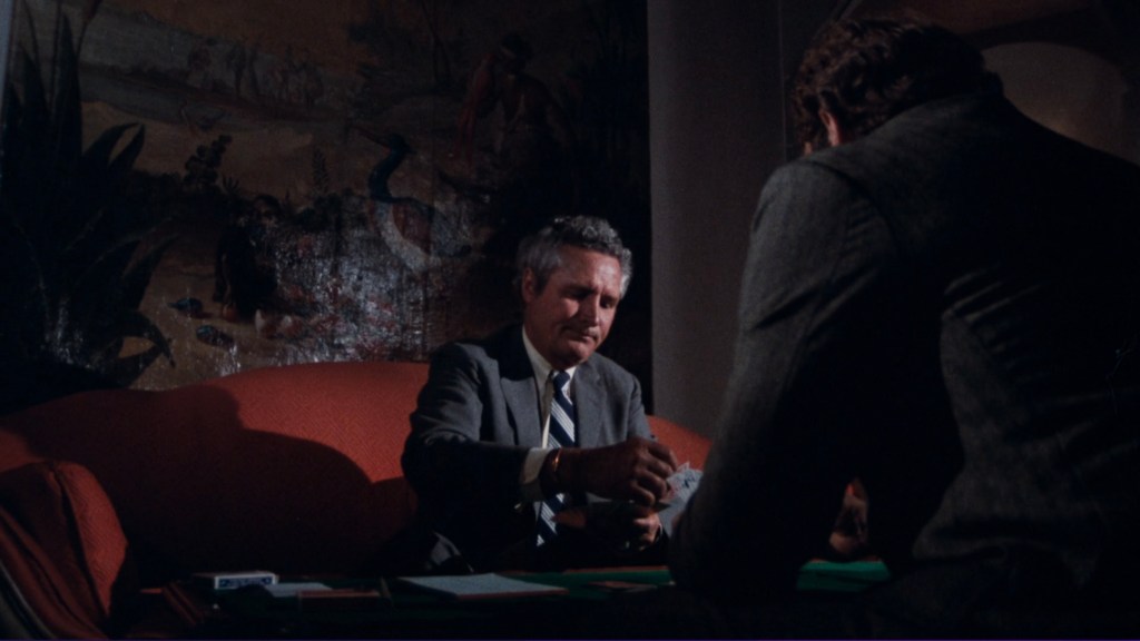 A man in a suit sits at a table, looking down as he deals cards, while another man in a grey jacket watches intently. The background features a painted mural with tropical themes.