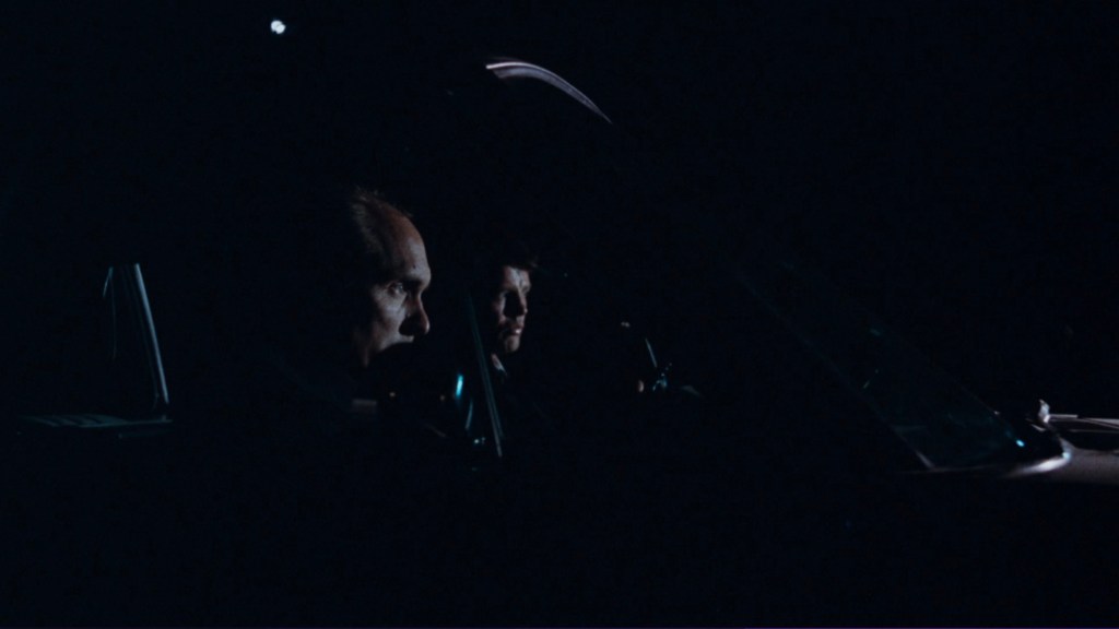 Two men sitting in a dark car, partially illuminated by the dashboard lights, with a focus on their serious expressions.