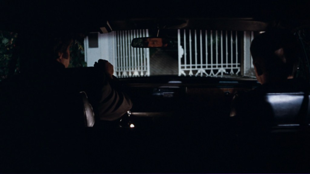 Two people sitting inside a car at night, looking ahead toward a house with a gated entrance.