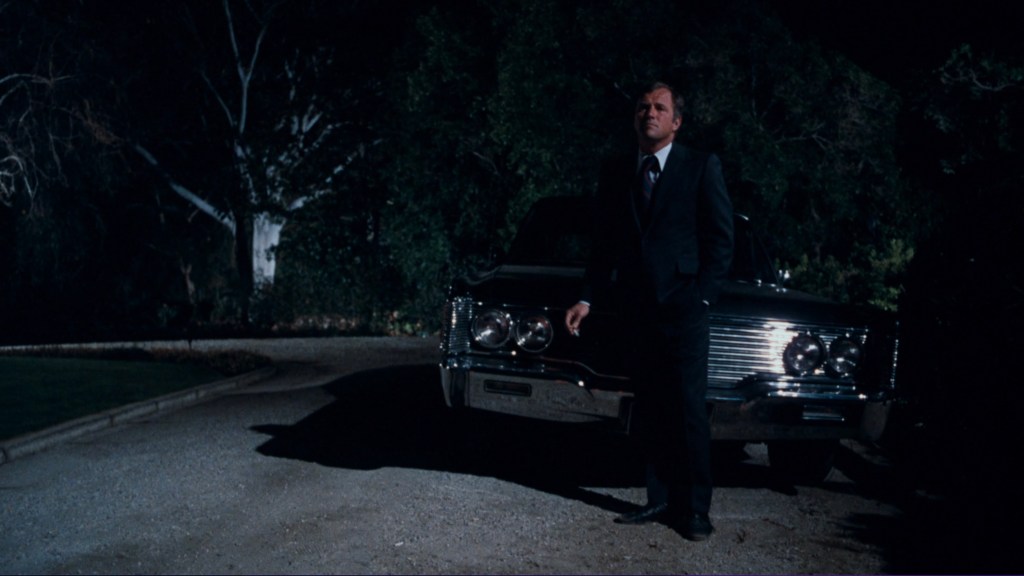 A man in a suit stands near a classic car at night, with shadowy trees in the background.