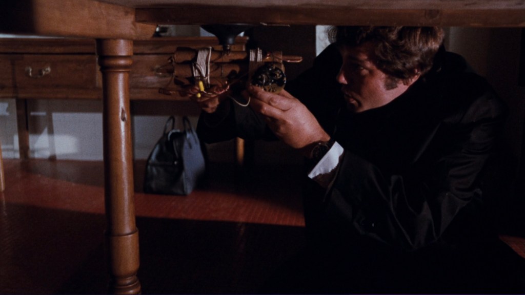 A man in a black suit inspecting a mechanical device under a wooden table.