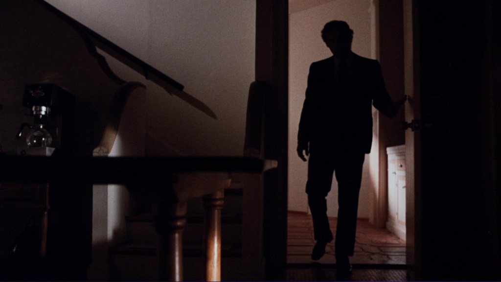 A silhouetted figure in a suit stands in a doorway, partially illuminated by light from the room behind, with a staircase and coffee maker visible in the background.
