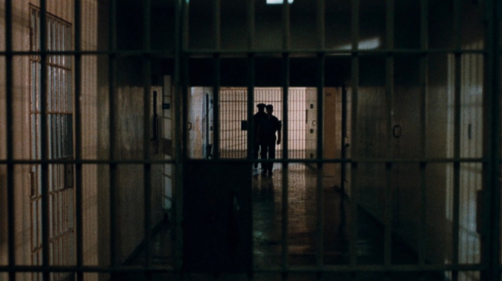 A dimly lit prison corridor with barred cells on either side and two figures walking in the distance.