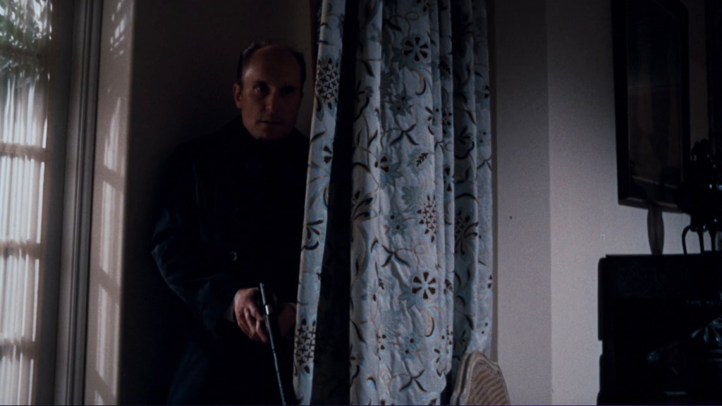 A man in a dark coat peeks from behind a floral curtain, holding a firearm, in a dimly lit room.