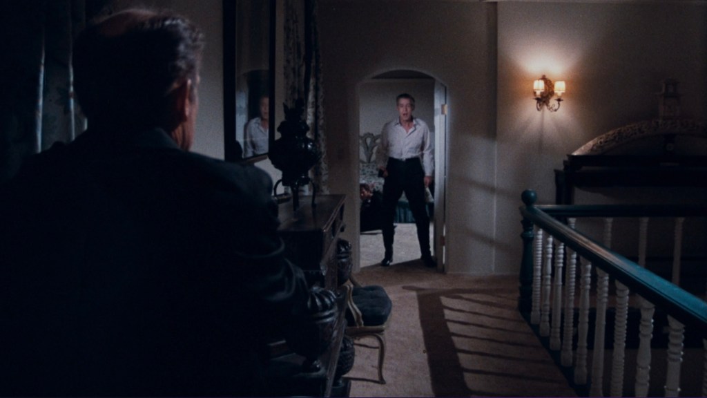 A scene showing a man seated, viewed from behind, gazing toward another man standing in a doorway, with soft lighting and a vintage interior design.