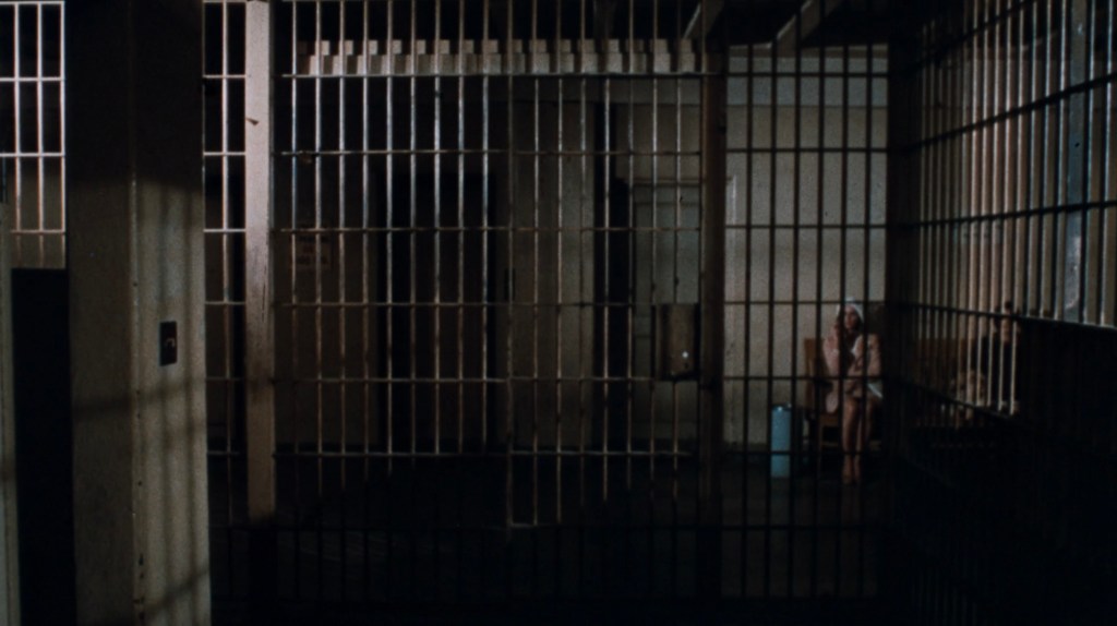 A dimly lit prison cell featuring metal bars, with a figure sitting in the corner.