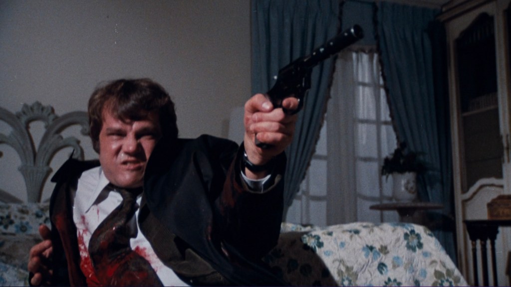 A man in a suit sits on a floral couch, holding a gun and appears to be in a tense situation. He has a pained expression and is covered in blood.