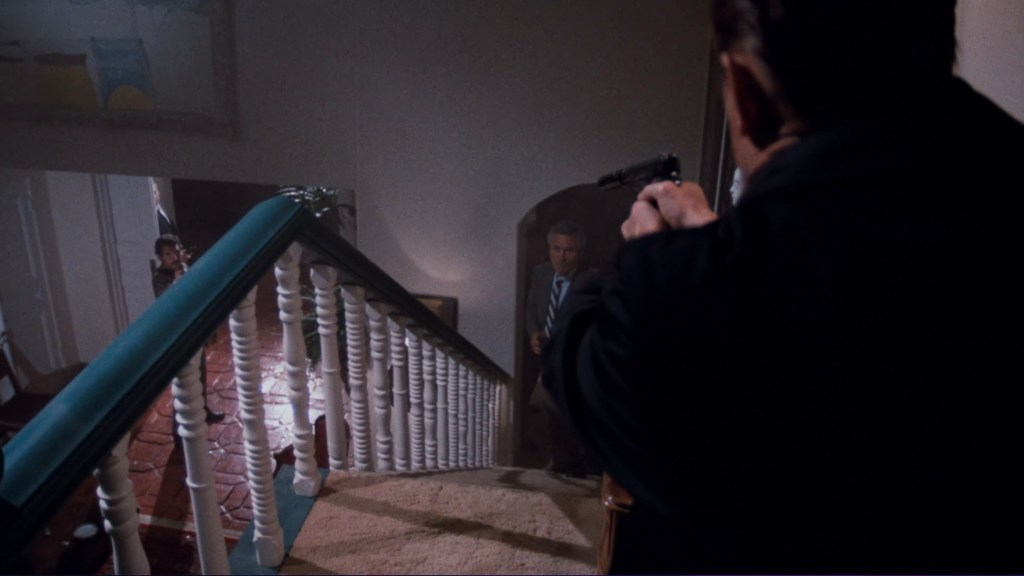 A dramatic scene showing a figure at the top of a staircase pointing a gun downwards, with a second figure visible at the bottom, creating a tense confrontation.