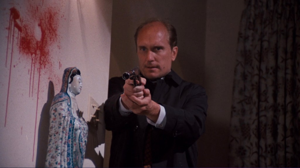 A man holding a handgun is pointing it towards the viewer, with a decorative porcelain figure beside him and red splatter marks on the wall.