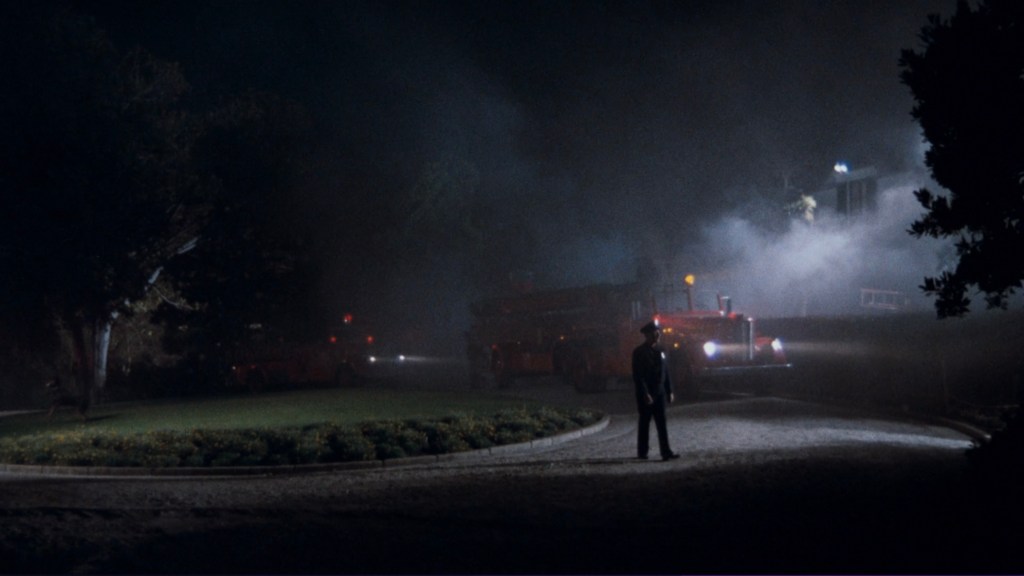 A police officer stands on a dark road surrounded by fog, with fire trucks visible in the background.