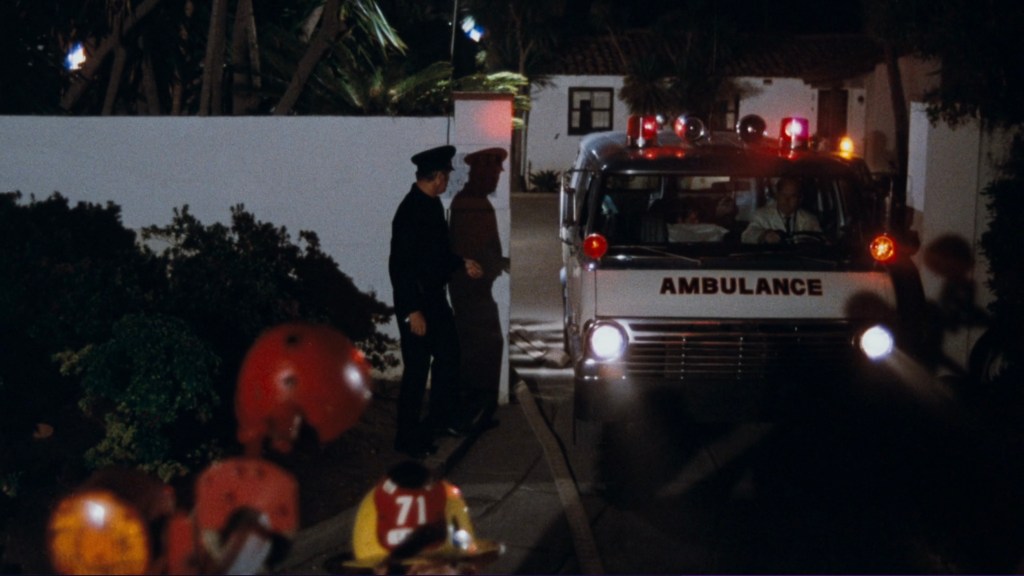 An ambulance with flashing lights is parked in a driveway at night, while a uniformed officer stands nearby.