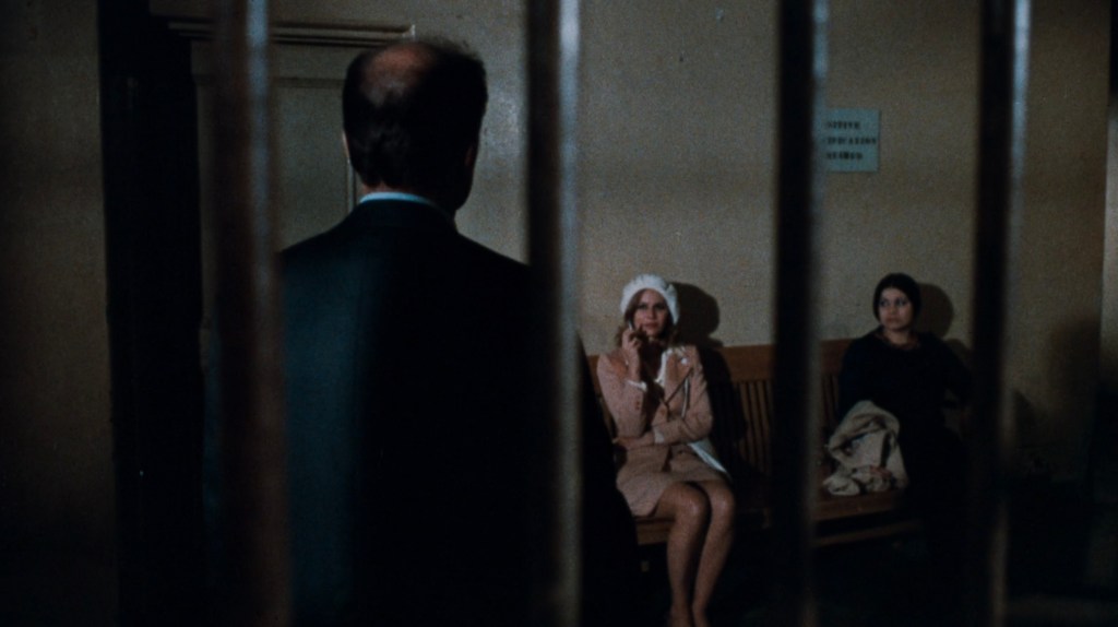 A scene depicting three figures in a dimly lit room, with a man in a suit facing away from the camera and two women seated on a bench, one wearing a light-colored coat and hat, and the other in dark clothing, separated by metal bars.