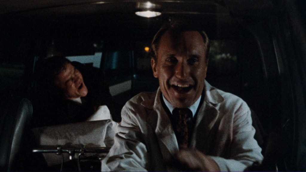 A man in a white lab coat smiles while driving a van, with a restrained individual visible in the background, appearing distressed.