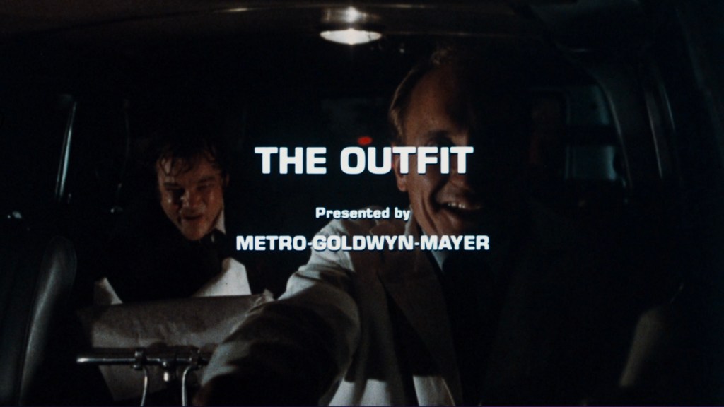 The movie title 'THE OUTFIT' displayed on a dark background inside a car, with two characters visible in the front seat.